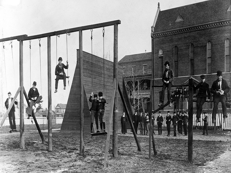 About a dozen young boys dressed in coats and hats are sitting on swings and jungle gyms in a dirt-lined playground.  A group of students, lay men and 2 Christian Brothers stand at the edge, in the back, and look on.  A group of 3 buildings are in the background.